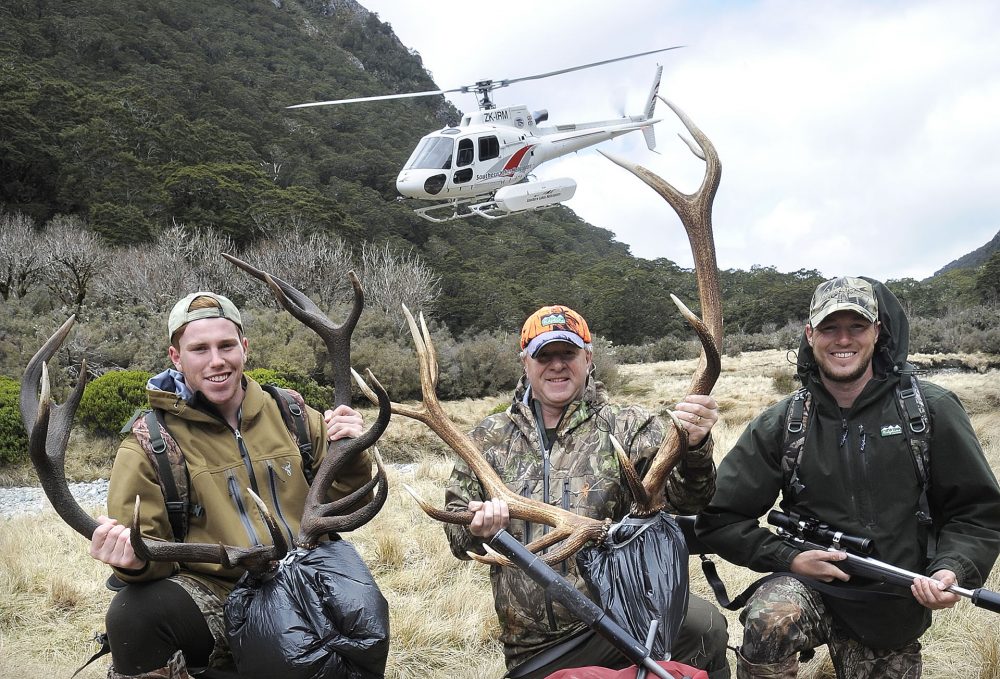 Three men standing with stags and a helicopter flying low in the background.